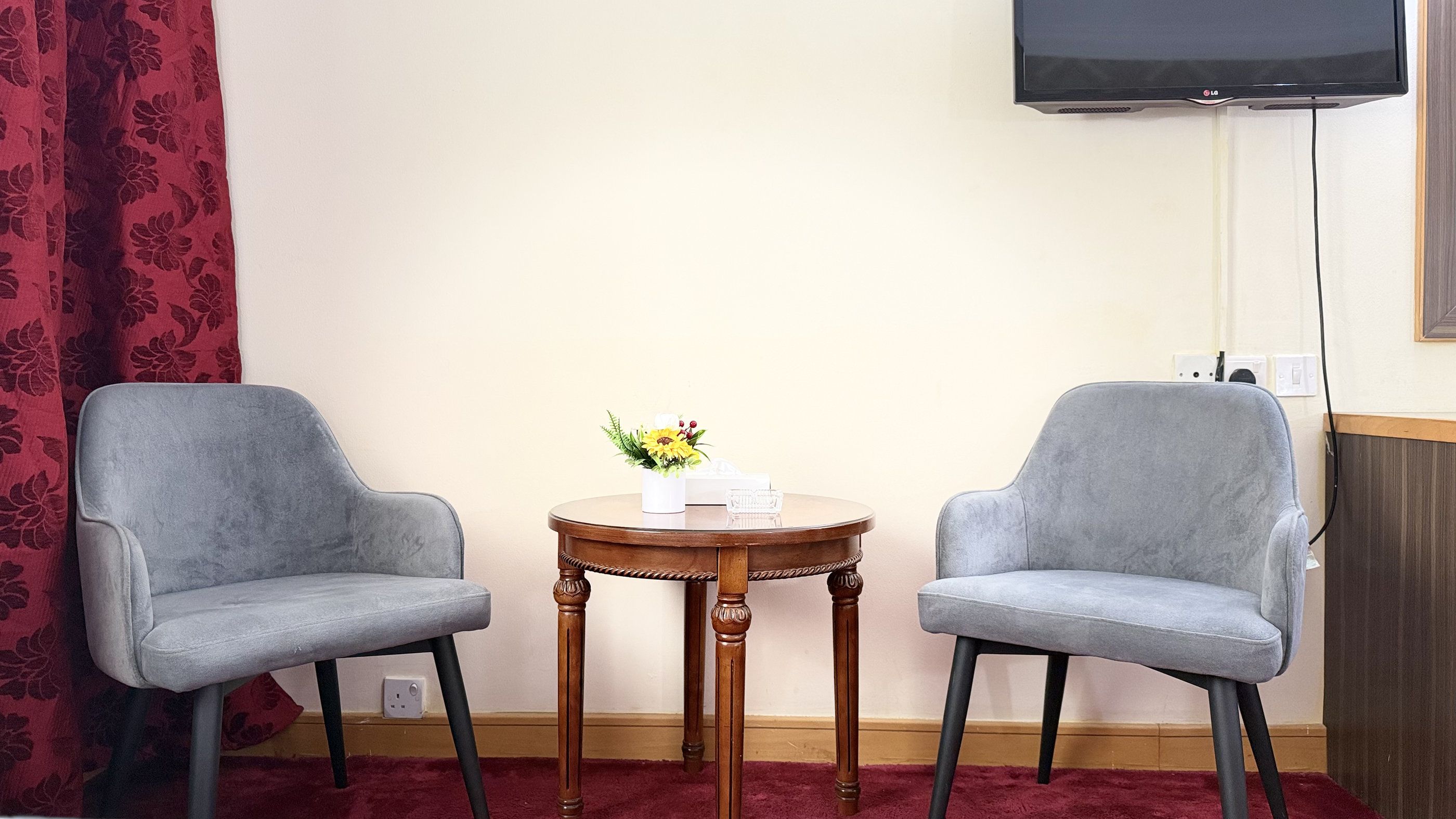 A room with two grey armchairs, a small wooden table with a vase of flowers, and a flat-screen TV mounted on the wall.
