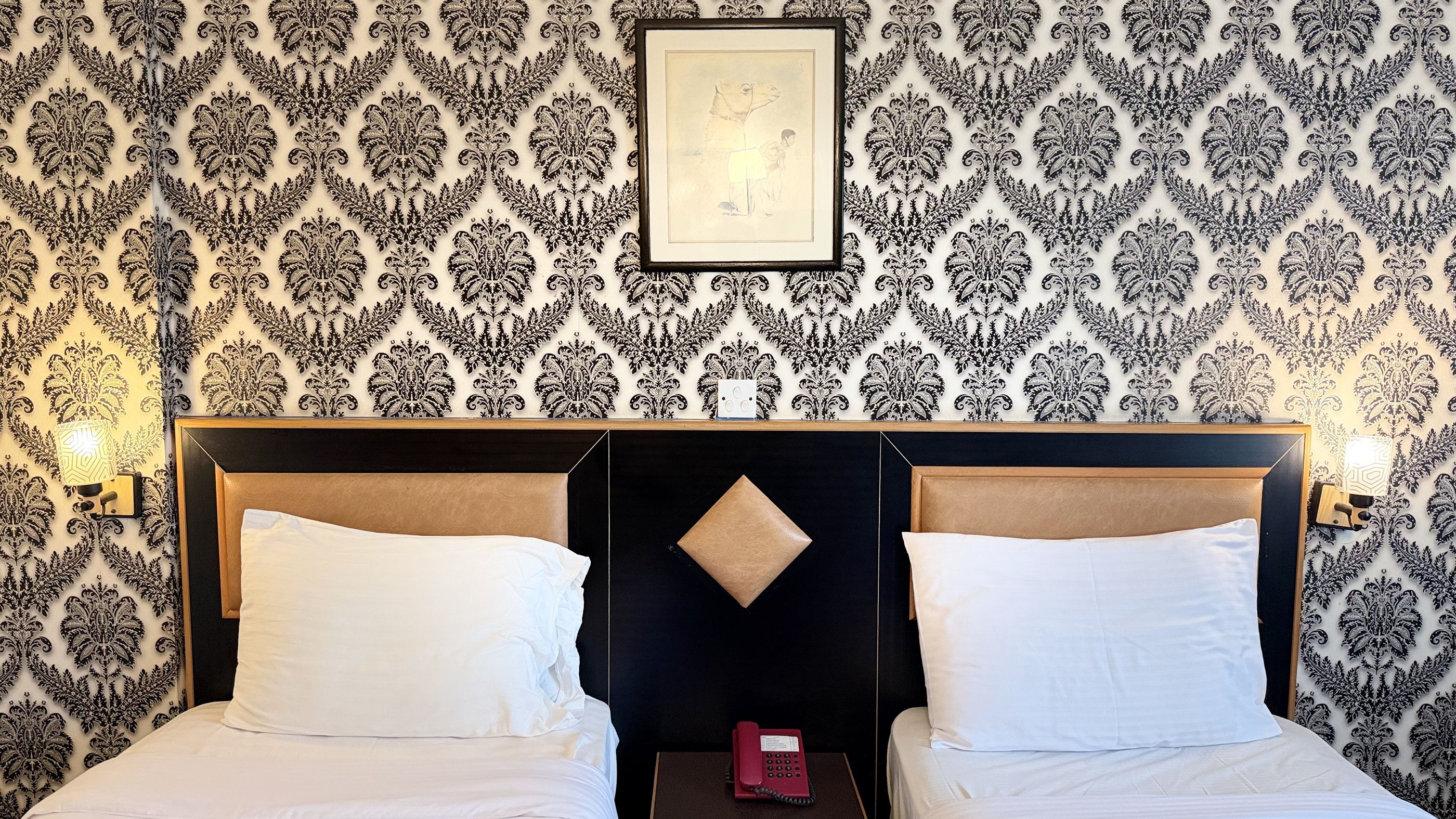 A hotel room with a patterned wallpaper, a framed artwork on the wall, and a bed with white linens.
