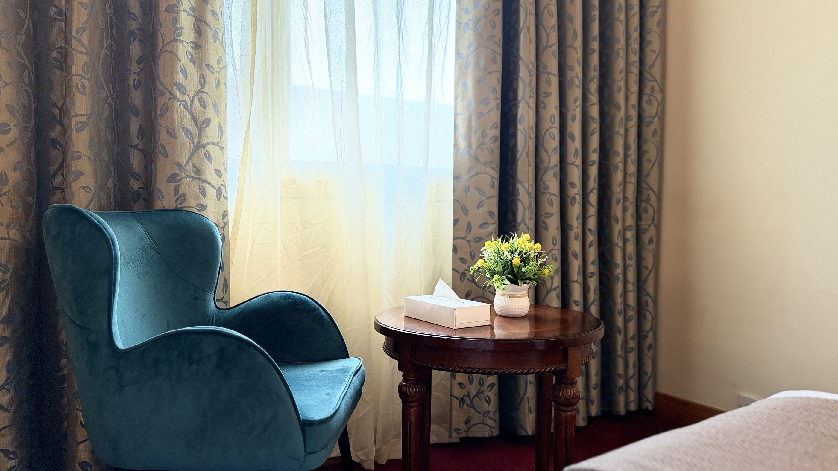 A room with a blue armchair, a small wooden table with a vase of flowers, a tissue box, and a bed with a white cover.