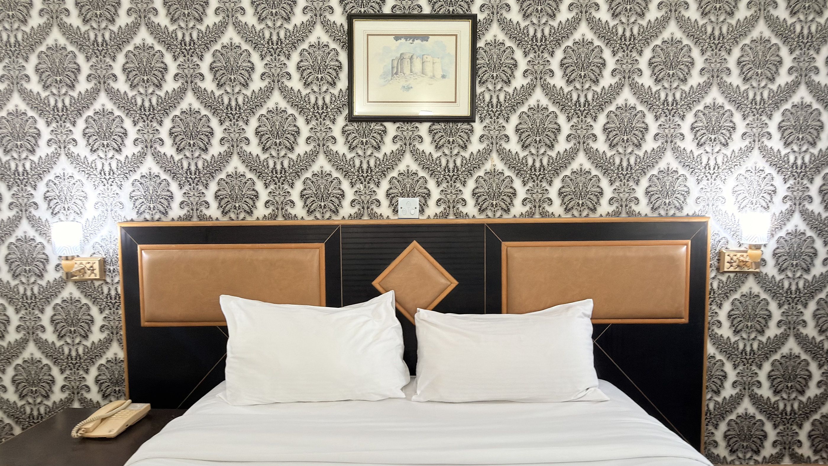 A hotel room with a patterned wallpaper, a bed with white bedding, and a framed picture on the wall.