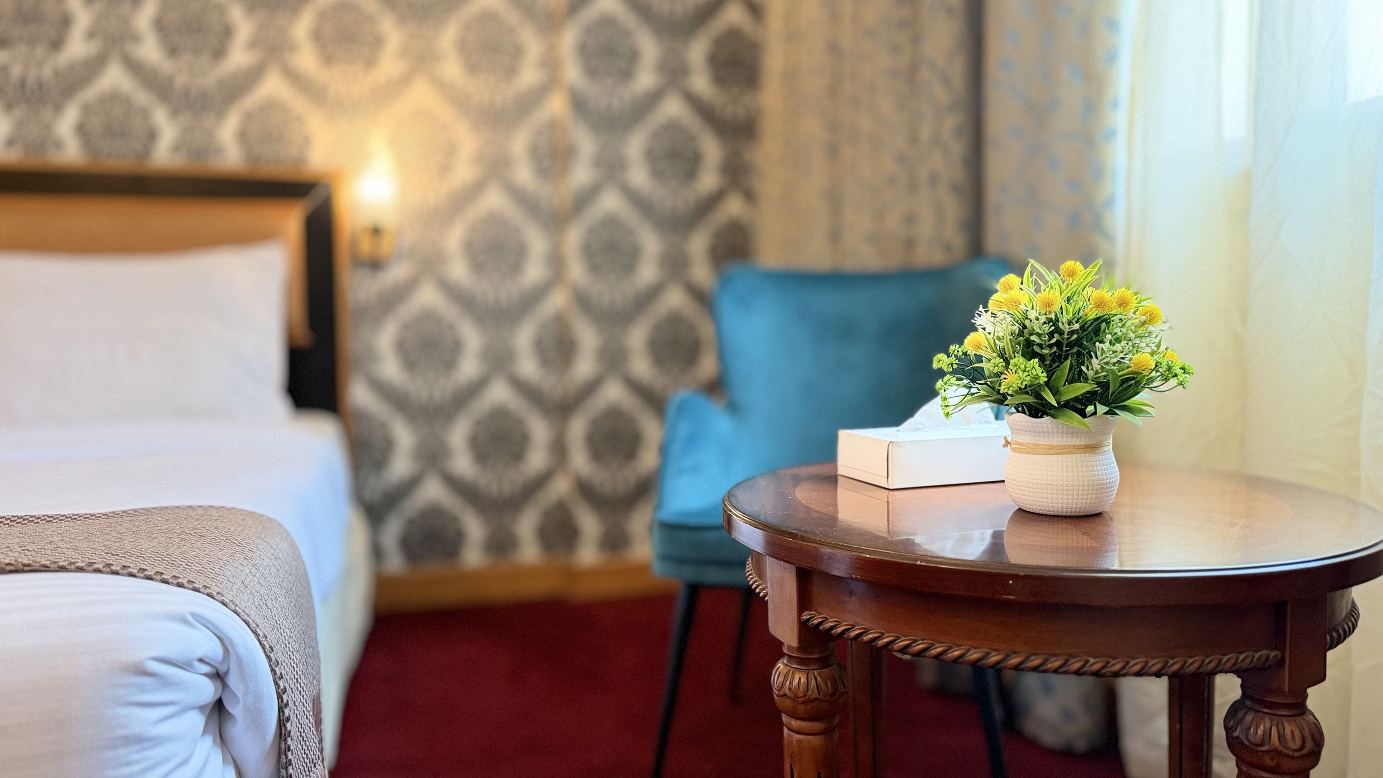 A hotel room with a bed, a small round table with a vase of flowers, a chair, and a patterned wallpaper.