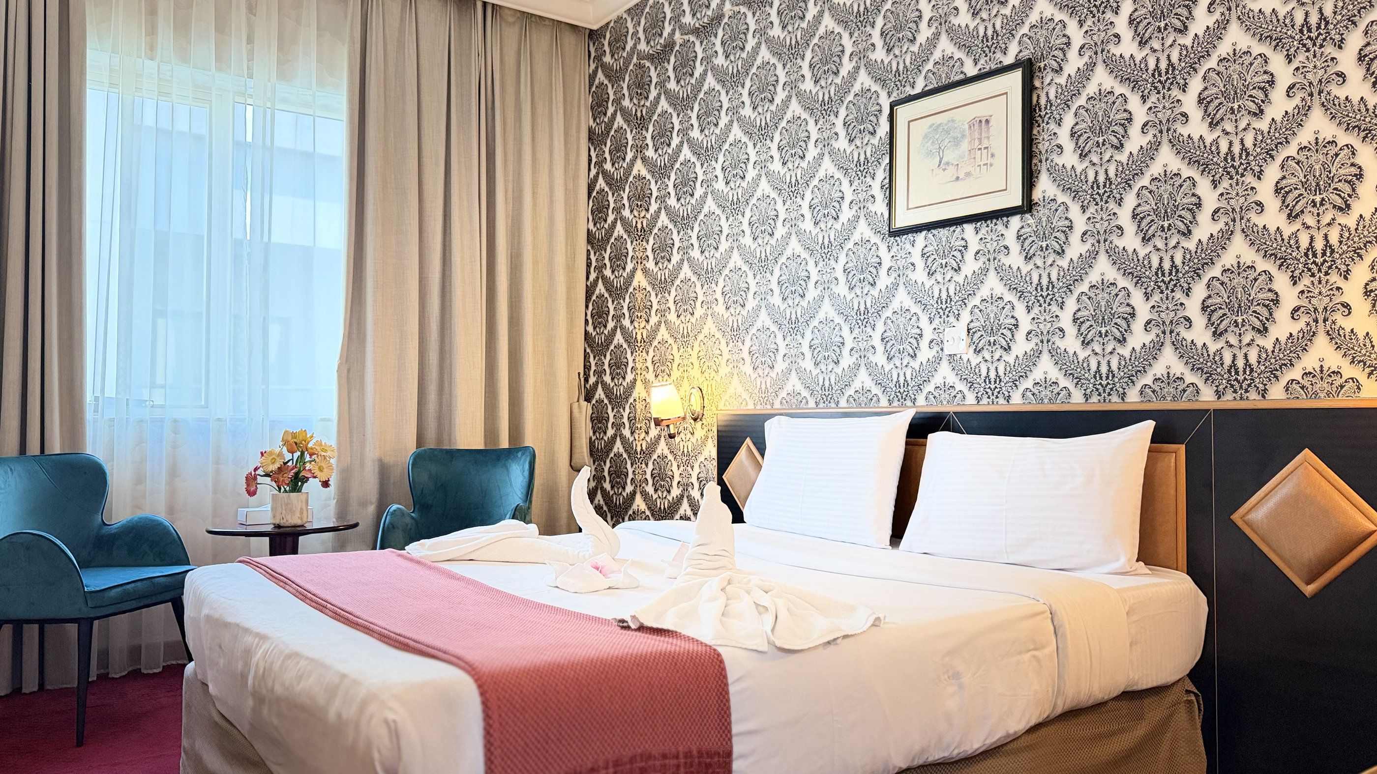 Une chambre d’hôtel avec un lit, deux chaises, une table de chevet, un vase avec des fleurs et un papier peint à motifs.