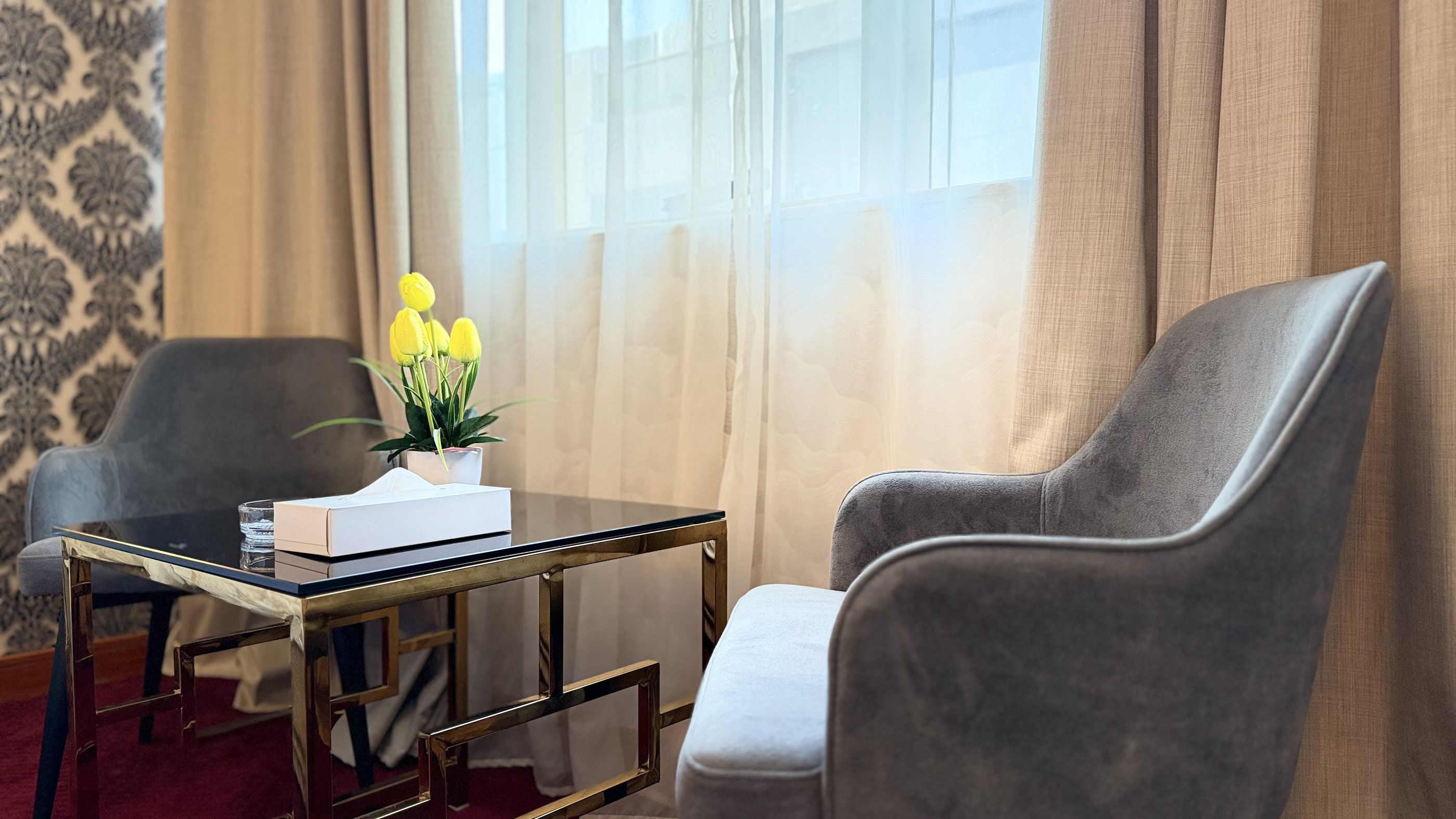Een hotelkamer met een grijze fauteuil, een bijzettafeltje met een vaas gele bloemen, en een raam met lichte gordijnen.