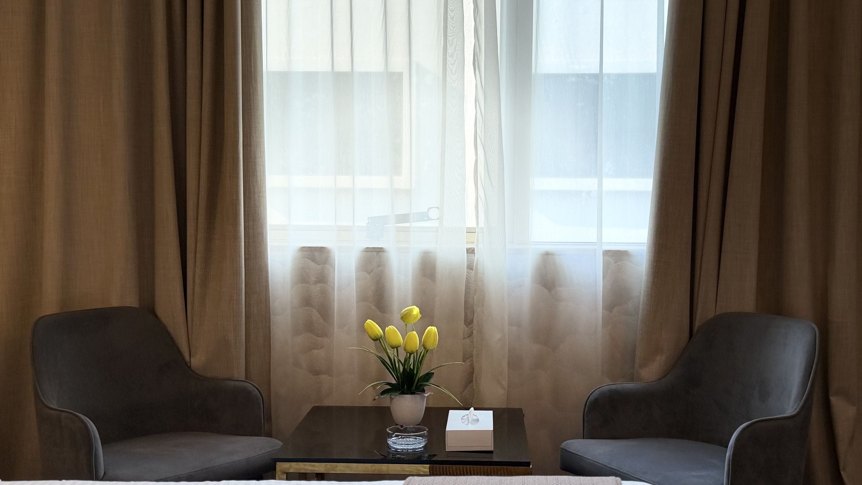 Een kamer met twee stoelen, een tafeltje met een vaas vol gele bloemen en een raam met dunne gordijnen.