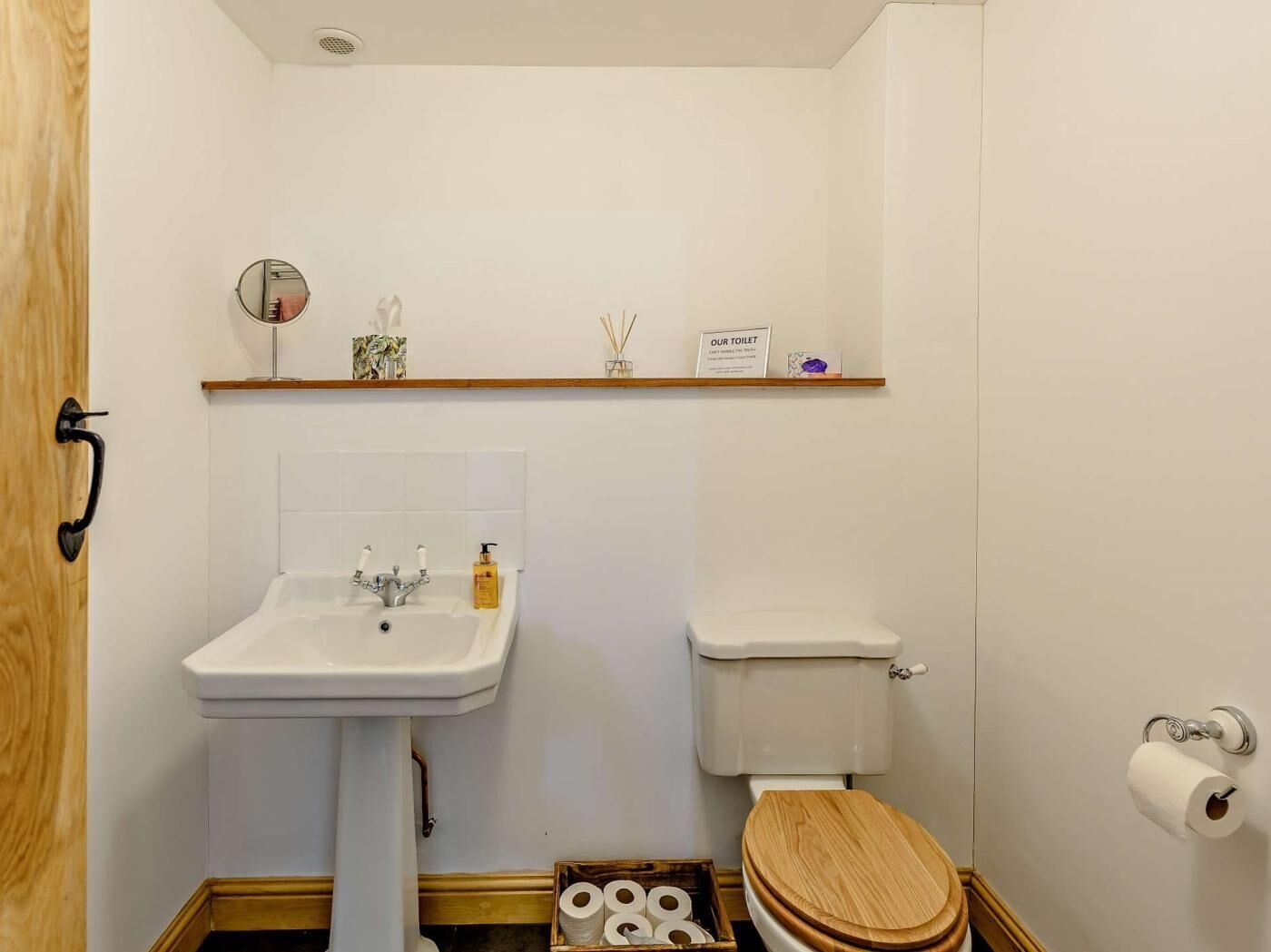 Une petite salle de bain avec une porte en bois, un lavabo blanc avec un miroir, des toilettes et une étagère au-dessus du lavabo.