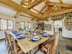 Un espace repas avec une longue table en bois dressée pour le repas, des murs en pierre et un plafond en bois.