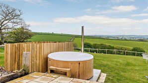 A wooden hot tub with a bench, surrounded by a wooden fence, on a stone patio with a view of green fields and hills. - 6 Bed in Torrington (oc-81519) (Great Torrington)