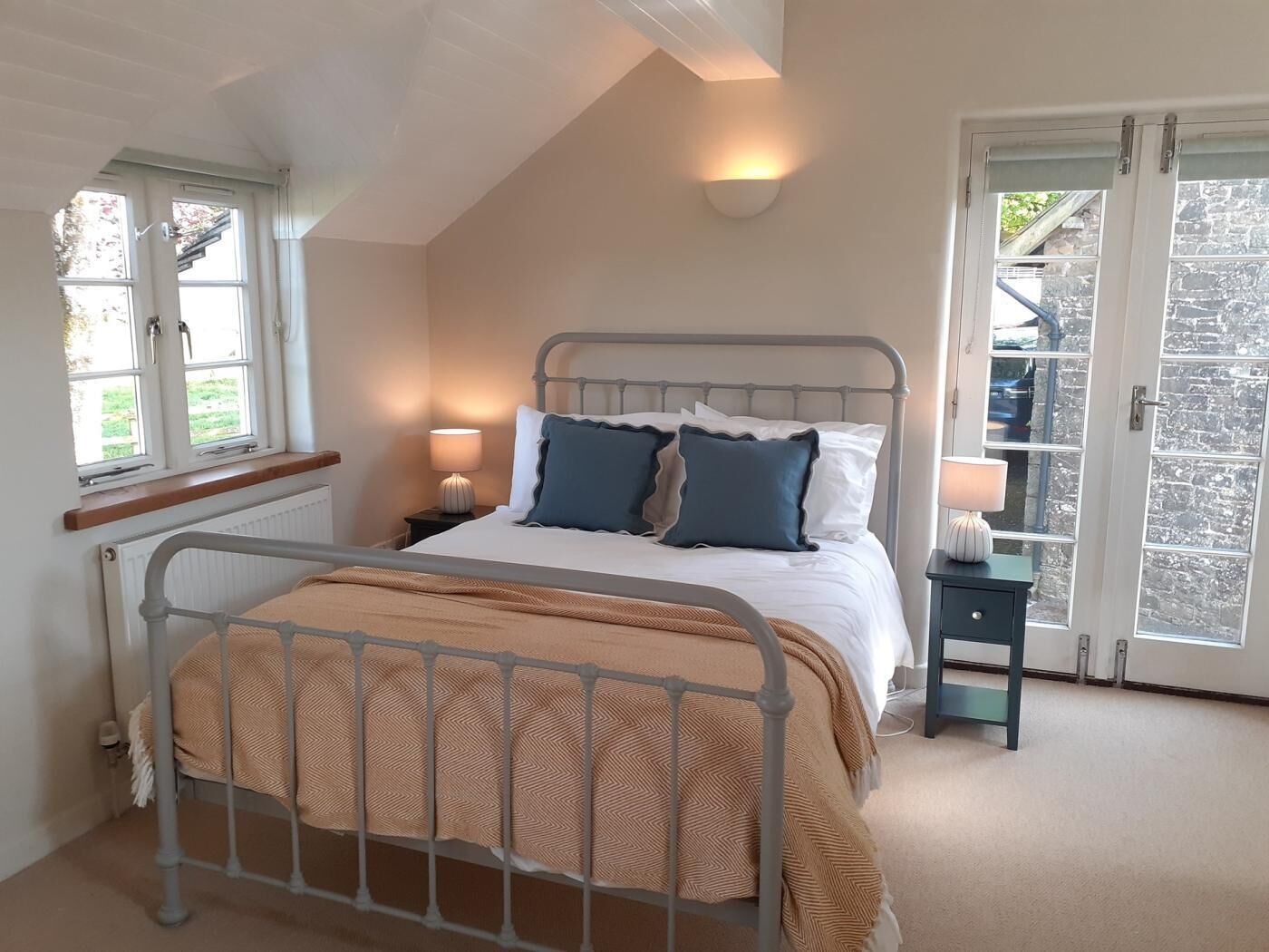 A bedroom with a metal bed frame, a window with a view, a bedside table with a lamp, and a door leading outside.