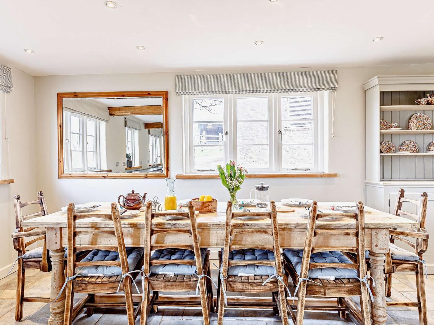 A dining room with a large table, wooden chairs with cushions, a mirror, and a window with curtains.