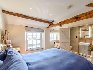 Ein Schlafzimmer mit einem großen Bett, einem Fenster, einem Badezimmer mit Spiegel und Waschbecken sowie Holzbalken an der Decke.