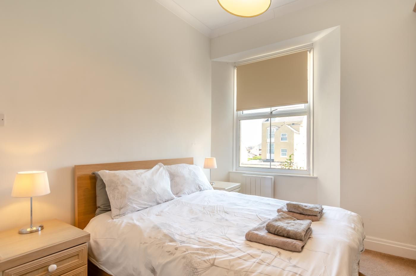 Een slaapkamer met een bed, twee bedlampen, een raam en uitzicht op gebouwen.