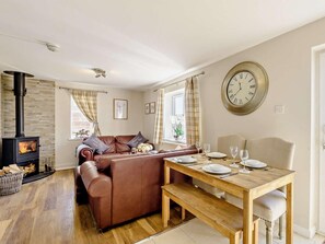 A cozy living room with a fireplace, a sofa, a dining table set for two, and a large wall clock. - 2 Bed in Holyhead (oc-81515) (Caergeiliog)