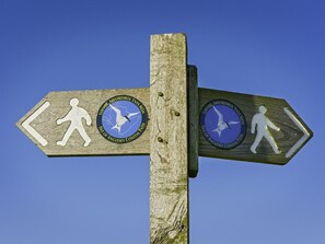A wooden directional signpost with two arrows, each featuring a walking figure and a circular emblem with text. - 2 Bed in Holyhead (oc-81515) (Caergeiliog)