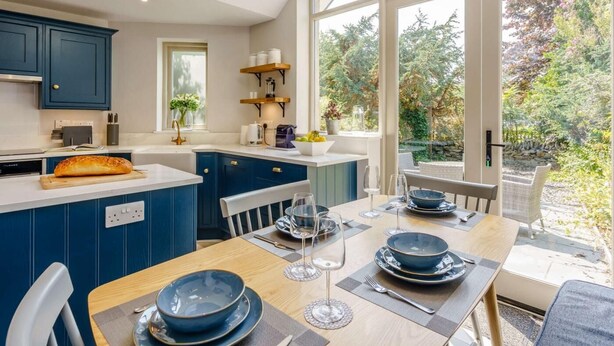 A modern kitchen with blue cabinets, a wooden dining table set for four, and a view of the garden through large windows. - 2 Bed in Grange-over-sands (oc-82672) (Cartmel)