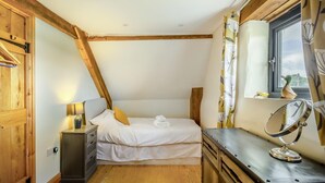 Ein Schlafzimmer mit schrägen Wänden, Holzbalken, einem Bett mit weißen Bettwäsche, einem Nachttisch mit Lampe, einer Kommode, einem Spiegel und einem Fenster mit Vorhängen.