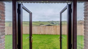 A view through a glass door with a wooden fence and a grassy yard. - 3 Bed in Ilfracombe (oc-82866) (Combe Martin)
