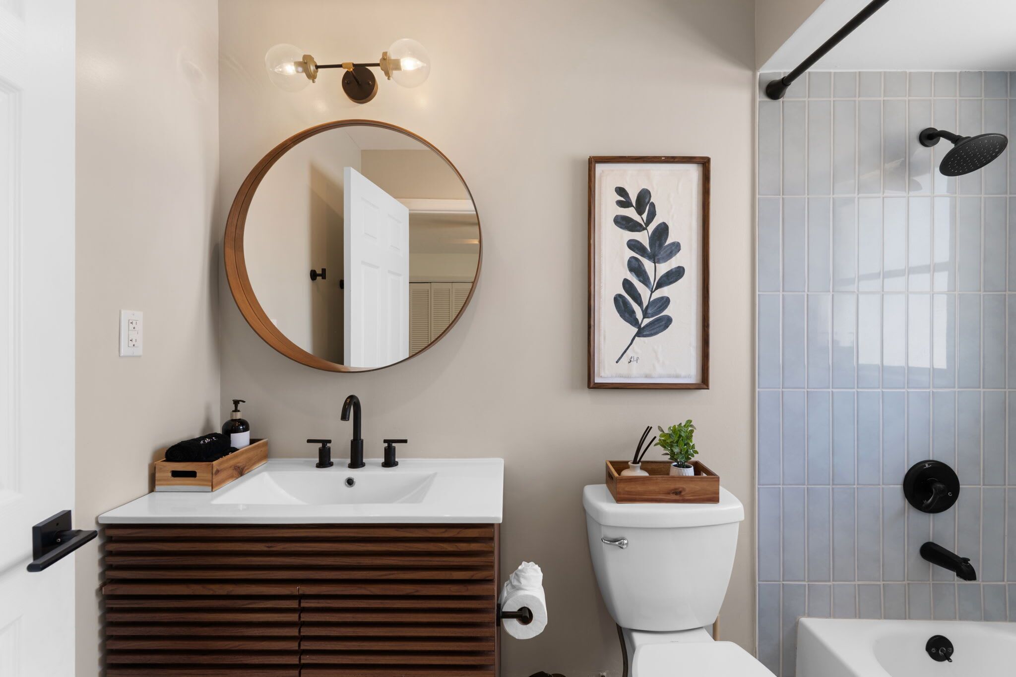 Une salle de bain avec un miroir rond, une œuvre d’art représentant une feuille encadrée, des toilettes et un espace douche.