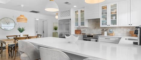 A modern kitchen with white cabinetry, a large island with bar stools, pendant lighting, and a stainless steel refrigerator.