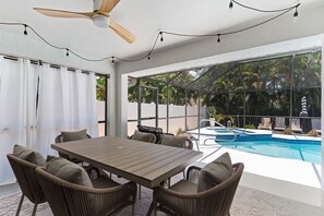 A modern outdoor patio with a wooden table and wicker chairs, a ceiling fan, and a view of a swimming pool. - Renovated Naples Home l Near Vanderbilt Beach (Naples)