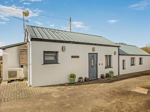 A single-story building with a metal roof, a small porch with potted plants, and a visible air conditioning unit. - 2 Bed in Beaworthy (oc-82819) (Ashwater)