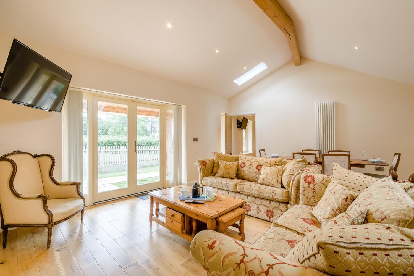 A spacious living room with a sofa set, a wooden coffee table, a television mounted on the wall, and a sliding glass door leading to an outdoor area.