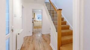 A hallway with wooden flooring, a staircase with a glass railing, and a kitchen visible through an open door. - 3 Bed in Torquay (oc-82959) (Torquay)