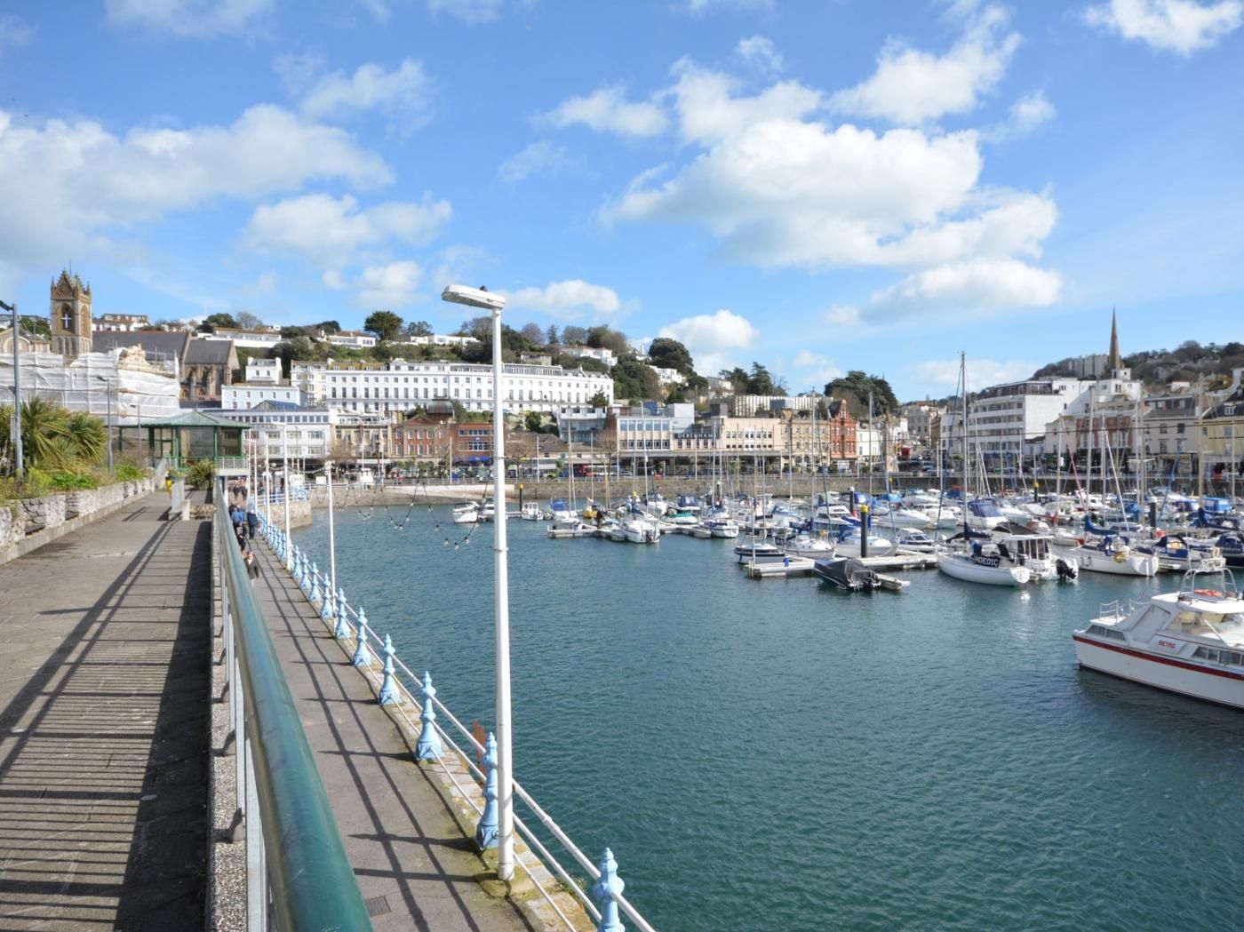 A marina with boats docked, a promenade, and buildings in the background.