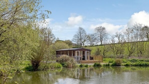A wooden cabin with a deck, surrounded by a pond and trees. - 2 Bed in South Brent (oc-82871) (South Brent)