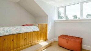 Quarto com teto inclinado, cama com colcha floral, um banco de madeira e janela com vista para árvores.