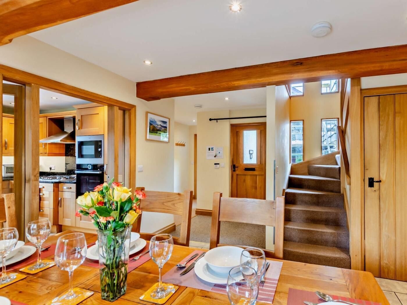 A dining area with a wooden table set for four, a kitchen in the background, and a wooden staircase.
