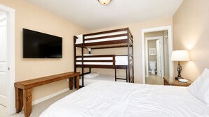 A bedroom with bunk beds, a flat-screen TV, a bedside lamp, and a wooden bench. - Pet Friendly Retreat - Outdoor Living - Sleeps 9 - Sunset Patios (Port Aransas)