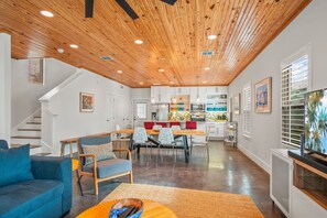 Ein modernes Interieur mit Holzbalkendecke, einer Essecke mit roten Stühlen, einer Küche mit weißen Schränken und einem Wohnzimmer mit blauem Sofa.