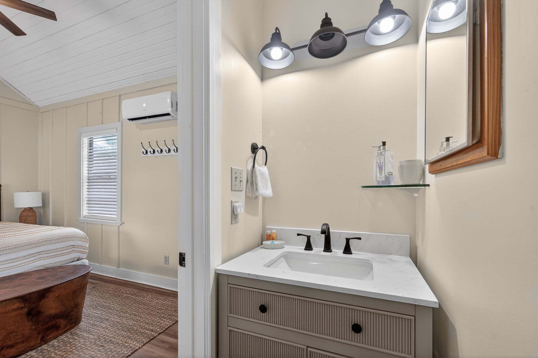 A bedroom with a bed, a wooden headboard, a window with blinds, an air conditioner, and a bathroom with a sink, mirror, towel rack, and shelf.