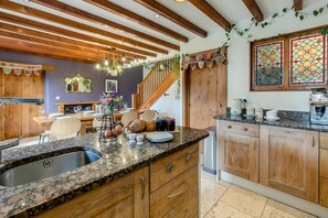 A kitchen with wooden cabinets, granite countertops, a coffee machine, and a dining area with a table and chairs. - 5 Bed in Truro (oc-tc82928) (Mount Hawke)