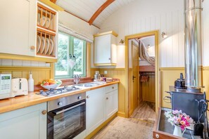 A compact kitchen with a gas stove, oven, and a small sink. There is a window with a view of greenery outside. - 1 Bed in Beaworthy (oc-82862) (Sheepwash)