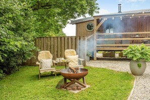 A backyard with a fire pit, two chairs, a wooden deck, and a small wooden structure with a chimney. - 1 Bed in Beaworthy (oc-82862) (Sheepwash)