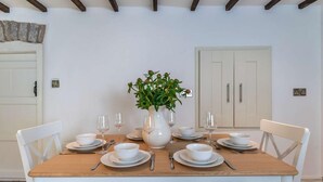 Une table à manger est dressée avec de l’assiette blanche, des bols et des verres à vin, et entourée de chaises blanches dotées de sièges rembourrés. Un vase blanc contenant des feuilles vertes est placé au centre de la table.