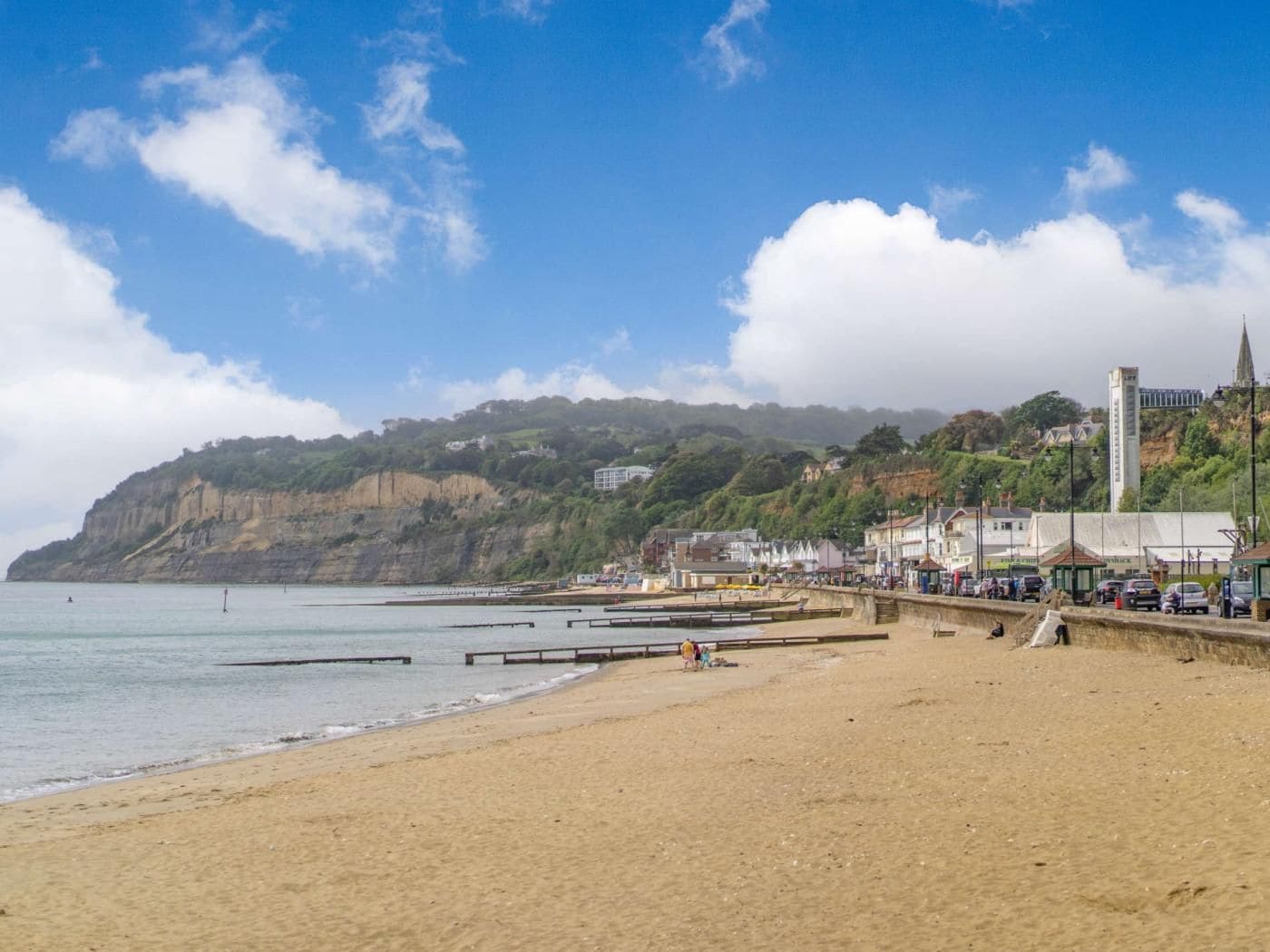 A beach with a promenade, a cliffside with buildings, and a church spire.
