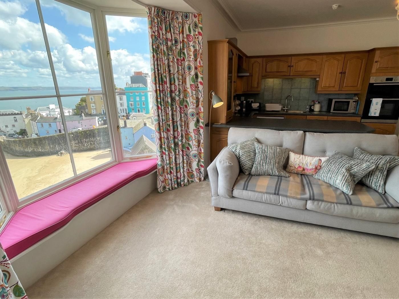 A living room with a sofa, a window with a view of buildings, and a kitchen area.