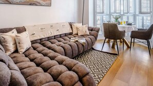 Un salon moderne avec un canapé d’angle, un coin repas avec une table et des chaises, et une grande fenêtre avec des volets.