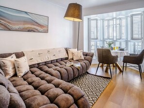 Ein modernes Wohnzimmer mit einer Eckcouch, einem Essbereich mit Tisch und Stühlen sowie einem großen Fenster mit Rollläden.