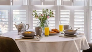 Une table de petit-déjeuner avec une bouilloire, une tasse, un bol, un verre de jus d’orange et un vase rempli de fleurs.