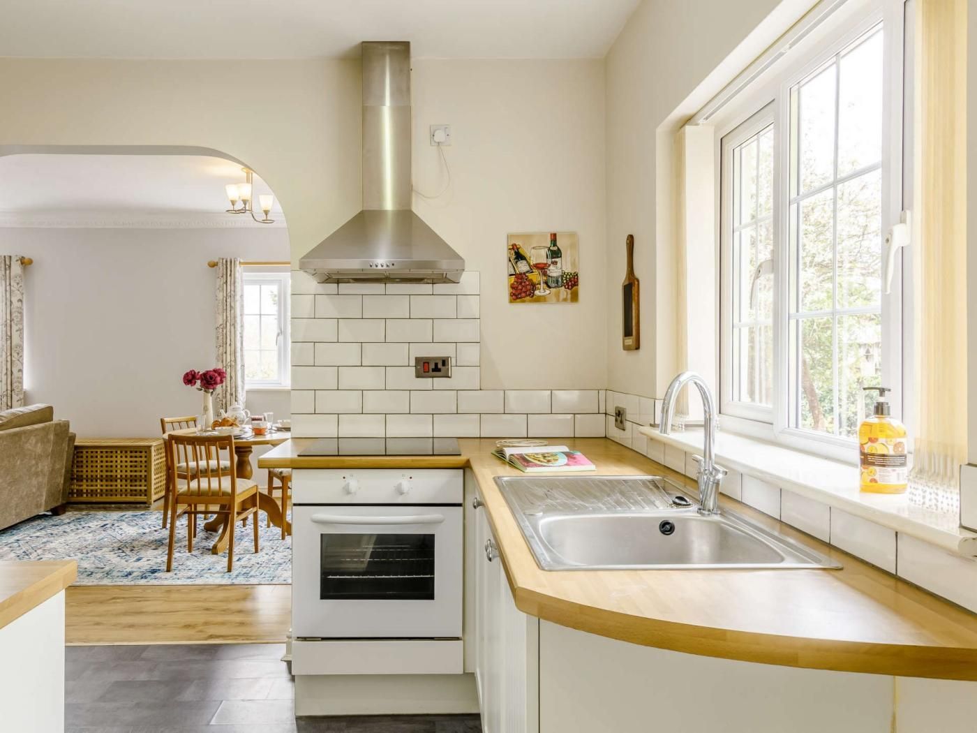 Una cocina moderna con armarios blancos, una campana extractora de acero inoxidable, horno integrado, encimera de madera y ventana con vistas a zonas verdes.
