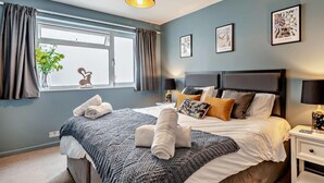 Ein Schlafzimmer mit einem Bett, einem Fenster, Vorhängen, einem Nachttisch und Wandbildern.
