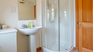 Un baño con ducha envuelta en vidrio, un lavabo blanco con artículos de aseo, y una puerta de madera.