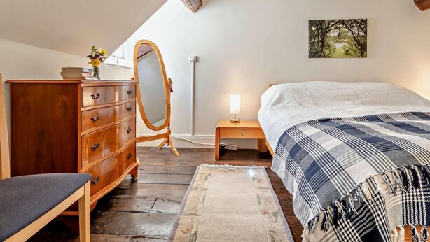 A bedroom with a wooden dresser, a mirror, a bedside table with a lamp, a bed with a patterned blanket, and a painting on the wall. - 1 Bed in Kidderminster (oc-82957) (Cleobury Mortimer)