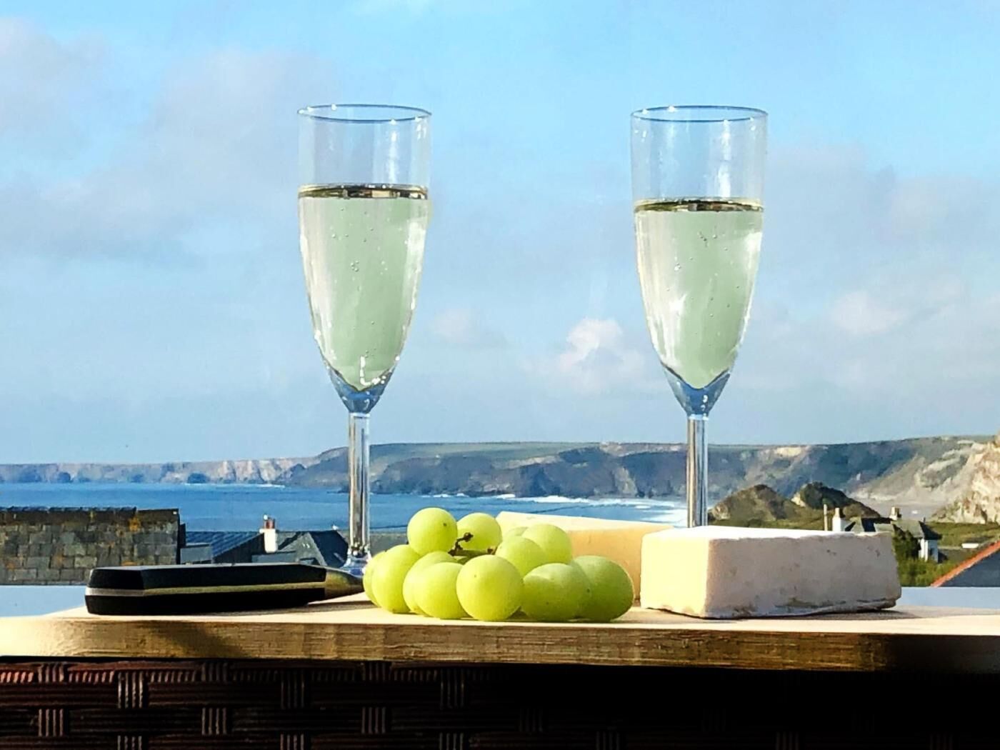 Zwei Gläser Weißwein, eine Traube grüner Weintrauben und ein Stück Käse auf einem Tisch mit Blick aufs Meer.