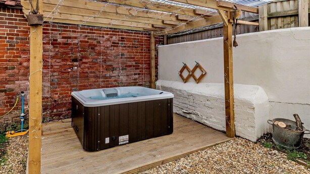 Un bain à remous sous une pergola en bois, dans un jardin avec un mur de briques et un mur blanc orné de motifs en forme de losanges.