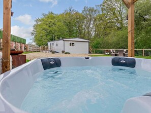 A hot tub with a wooden structure in the foreground, a small house in the background, and a fenced area with trees.