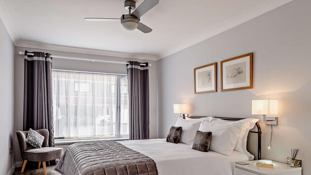 Een hotelkamer met een bed, nachtkastje, stoel, plafondventilator en raam met gordijnen.